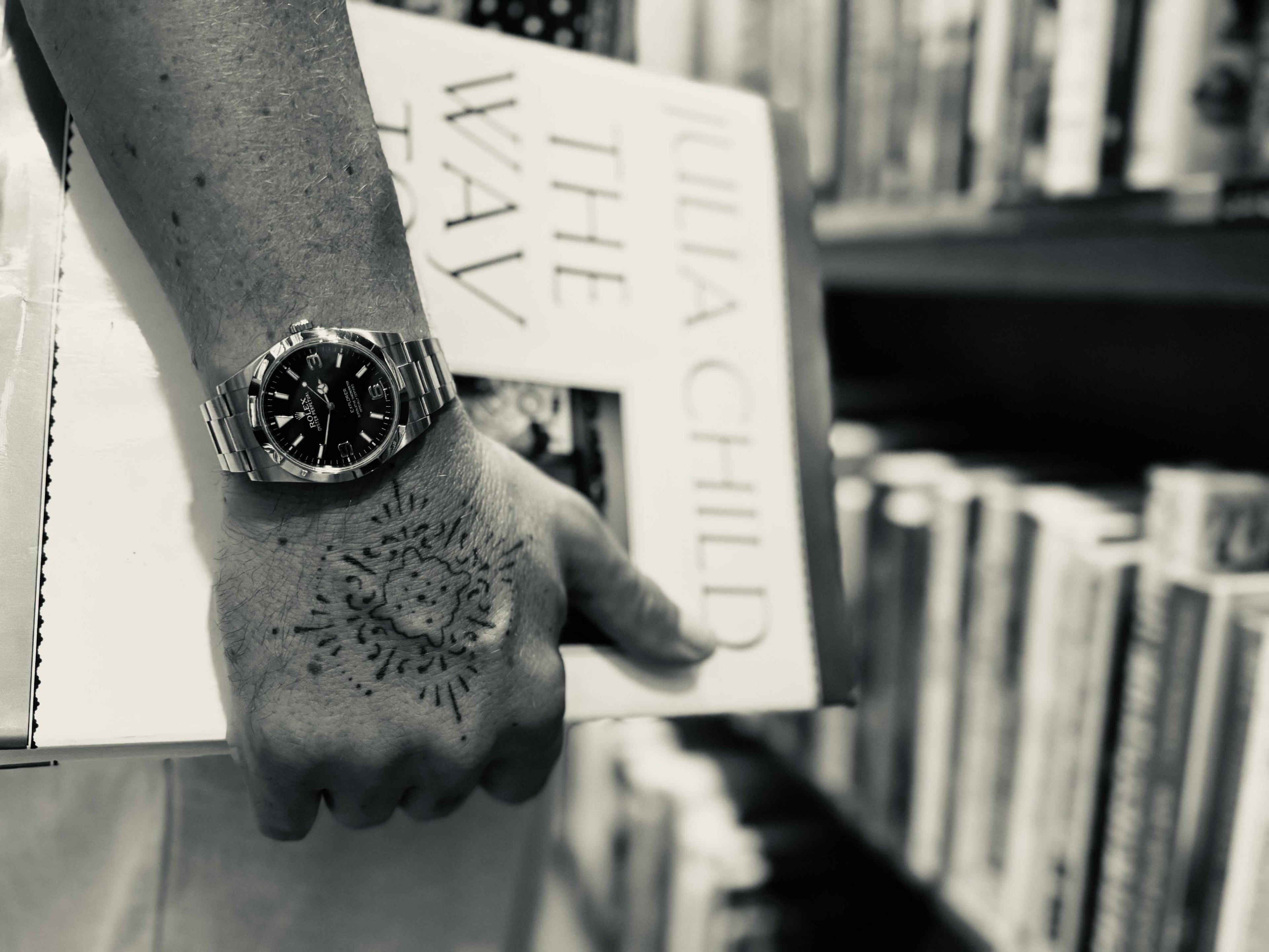 Hand holding a book with a tattoo on the palm, wearing a watch, against a blurred bookshelf background.